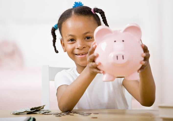 A young girl wearing a white shirt is holding up a pink piggy bank.
