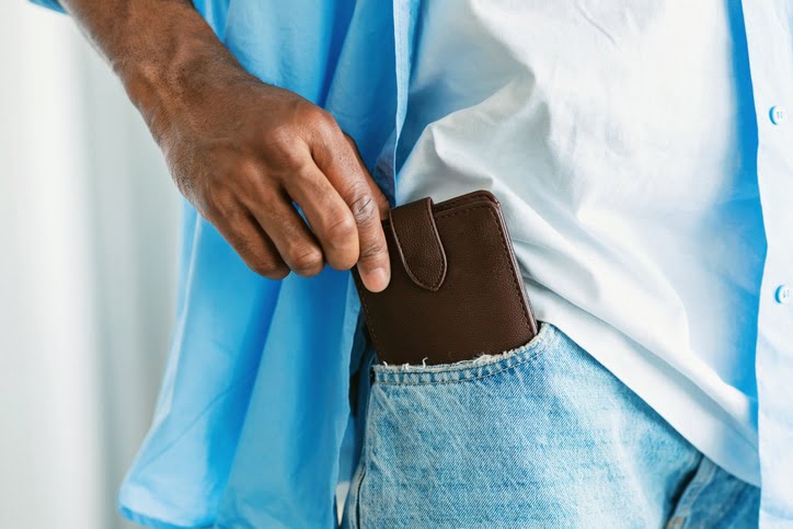 The hand of a man in a blue button-up shirt over a white t-shirt puts a brown leather wallet into the pocket of his jeans.